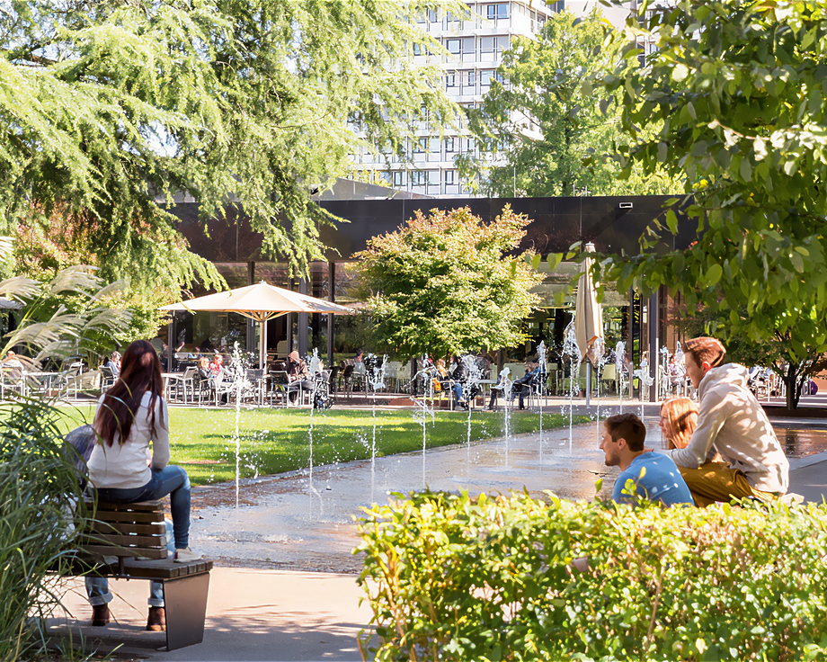 Menschen sitzen auf Bänken neben einem Brunnen mit Wasserspielen, im Hintergrund Café mit Gästen und Bäumen.