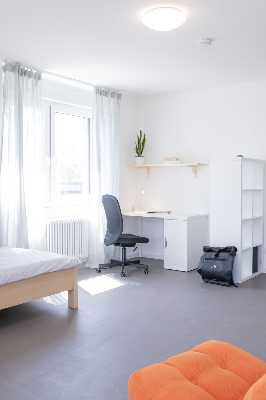 Ein helles Zimmer mit Bett, Schreibtisch, Stuhl, orange Sessel, Regal und Fenster mit Vorhang. Deko zeigt eine Stadt-Silhouette, daneben Spiegel und Pflanze.